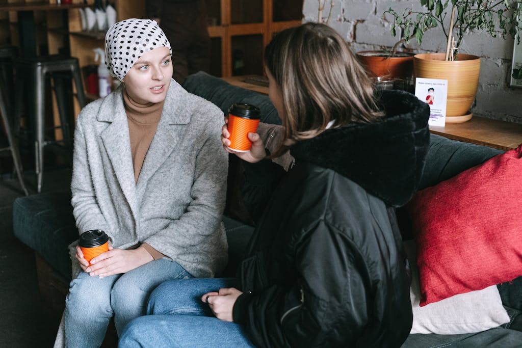Two women in winter clothing chatting over coffee on a couch indoors.