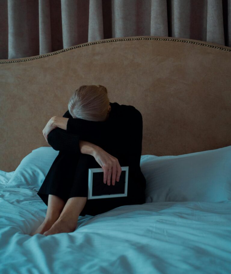 A senior woman sits on a bed holding a photo frame, expressing loneliness and grief.