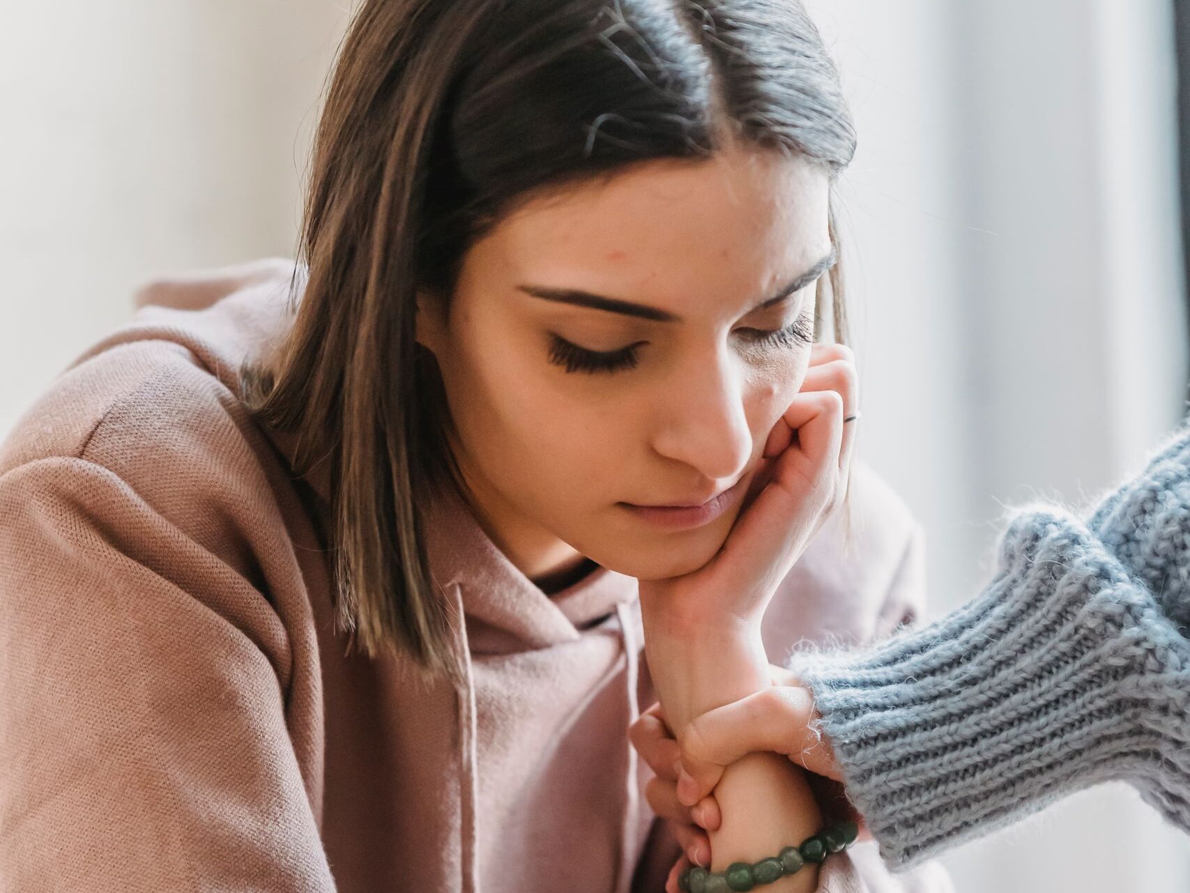 Faceless female grabbing hand of unhappy pensive girlfriend sitting in light living room and looking down during quarrel at home