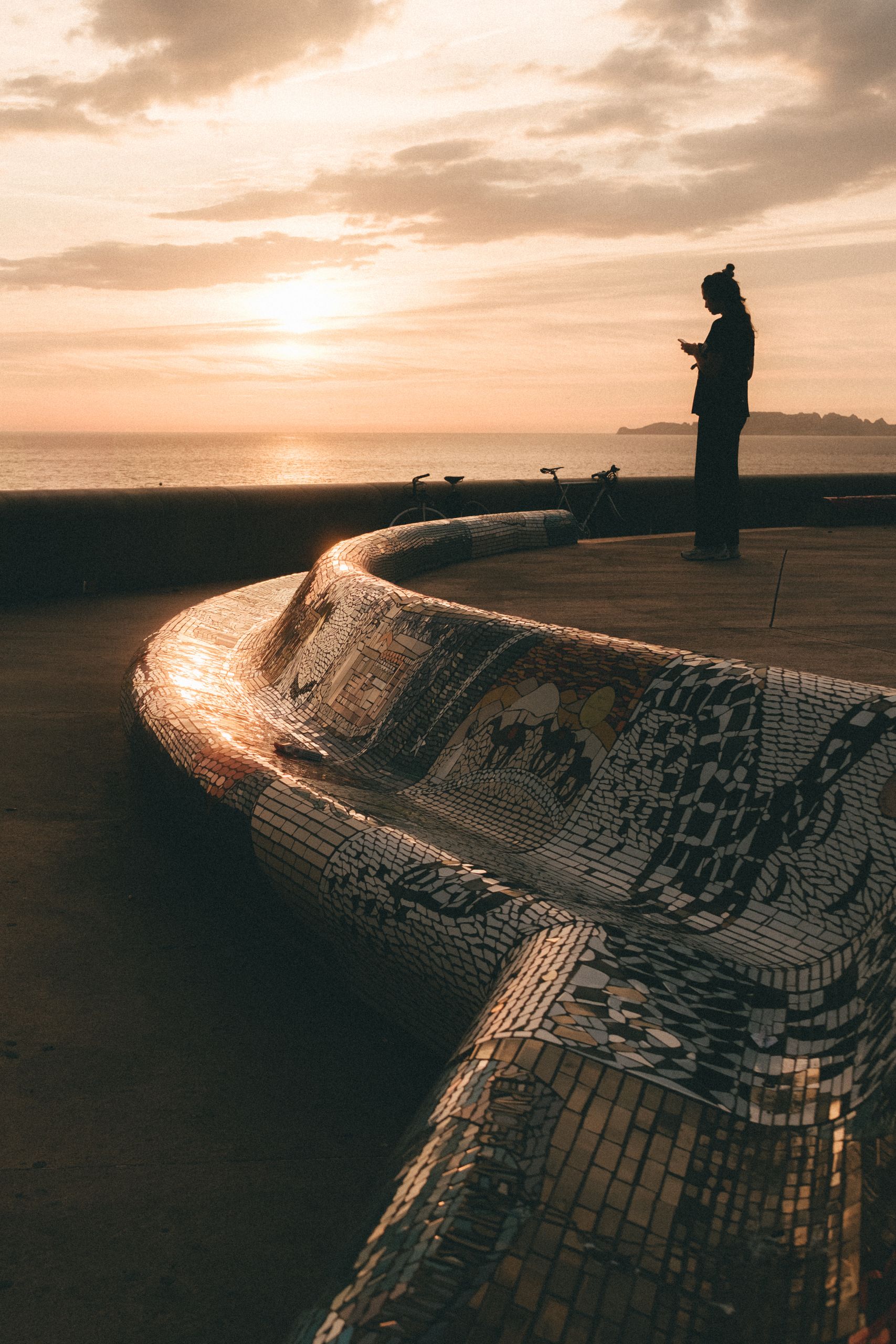 Person silhouetted against ocean sunset on an artistic mosaic bench, capturing serene waterfront view.