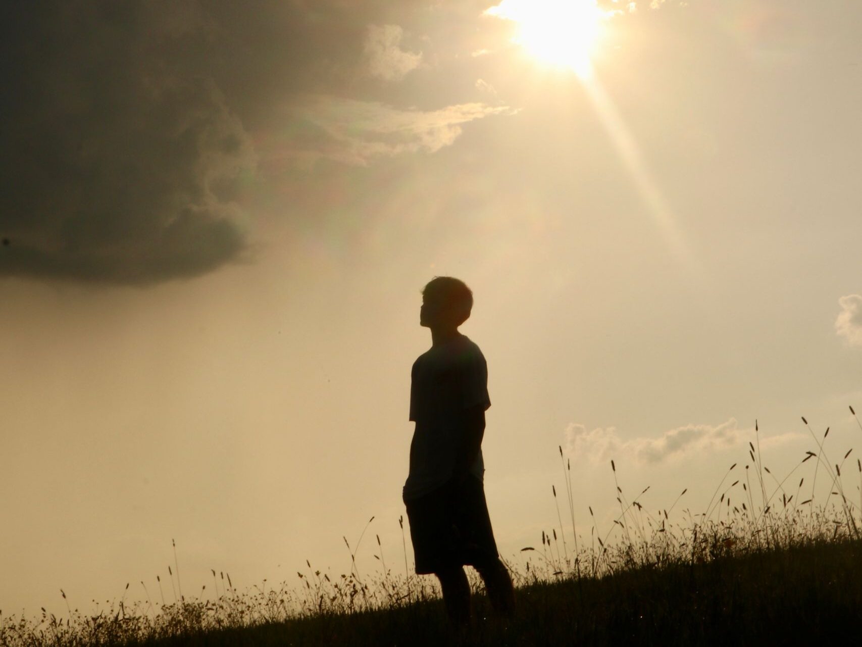 Silhouette of an individual on a grassy hill beneath a dramatic cloudy sky with sunflare.